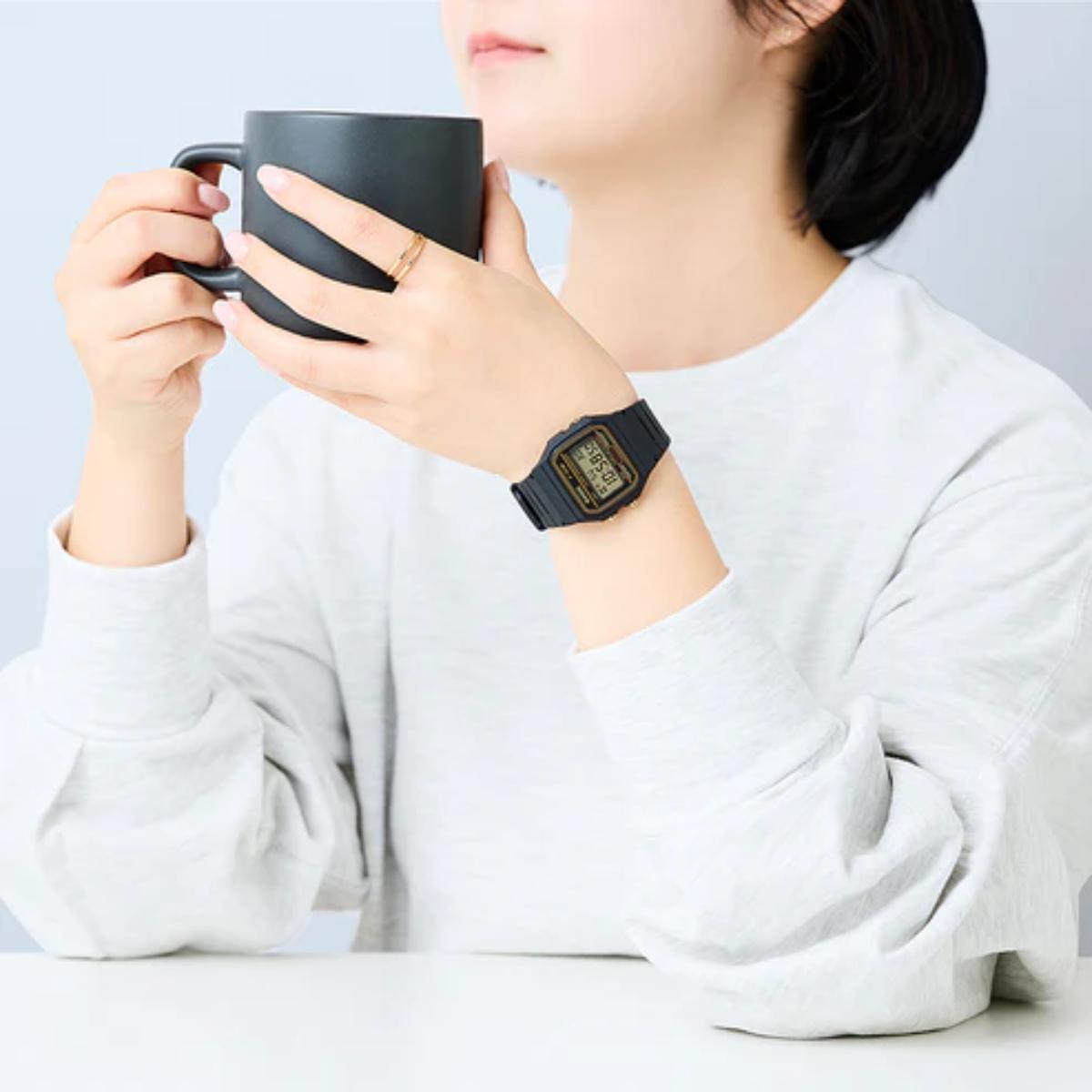 Person holding a black mug with a watch on wrist, wearing a white shirt against a light gray background