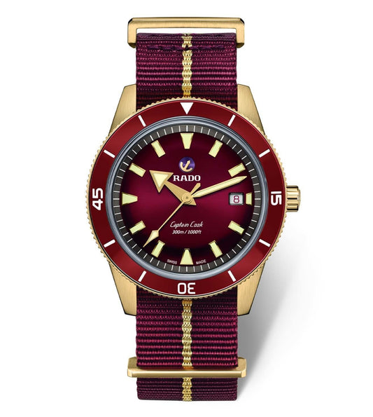 A Rado Captain Cook Automatic bronze watch with a red dial, gold case, and a red and white striped strap.