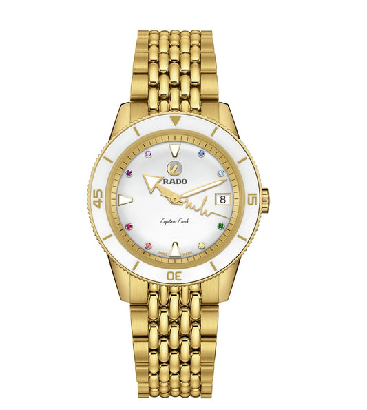 A gold-colored Rado Captain Cook Marina Hoermanseder Heartbeat watch with a white dial, multicolored stones on the bezel, and a gold bracelet.