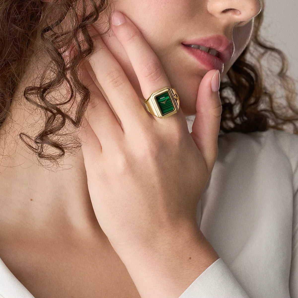 Close-up of a person wearing a gold ring with a green gemstone, touching their face.