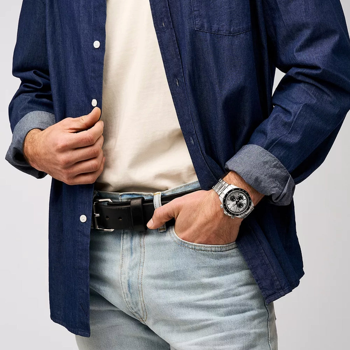 Person wearing a blue denim shirt, white t-shirt, light blue jeans, and a watch on a plain background