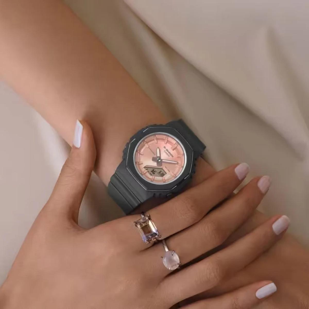 Black watch with pink face on a wrist, wearing two rings.