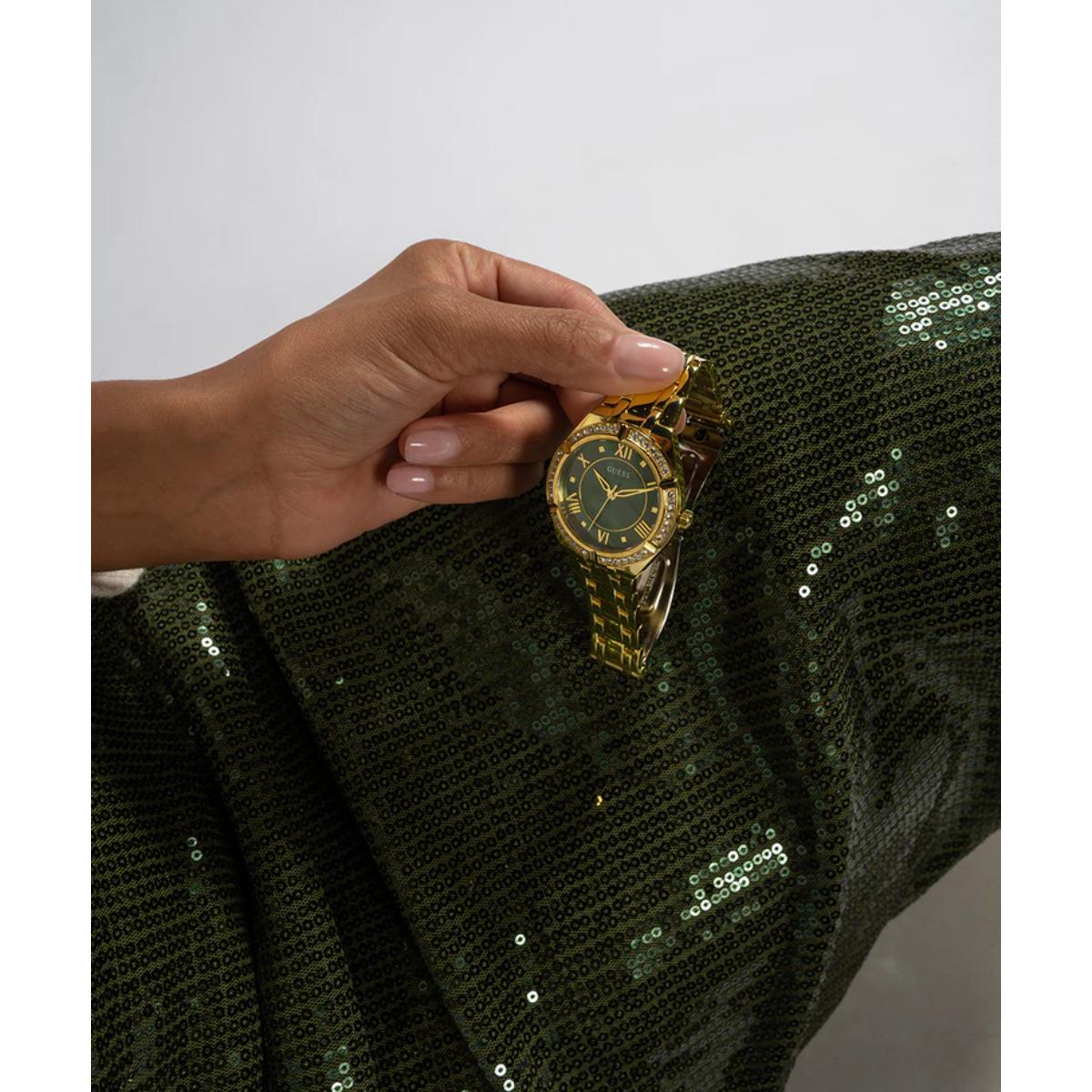 Hand holding a gold watch with a green face on a green sequined fabric background