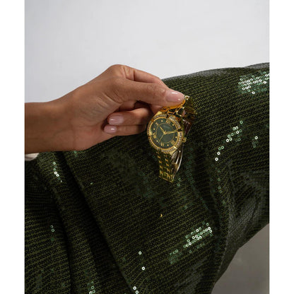 Hand holding a gold watch with a green face on a green sequined fabric background