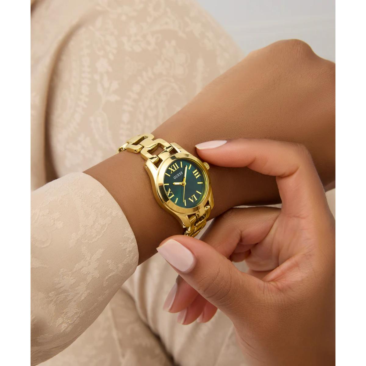 A close-up lifestyle shot of a woman wearing a gold-tone watch with a green dial and Roman numeral hour markers. Her hand is adjusting the strap, and she is wearing a beige lace-trimmed top.