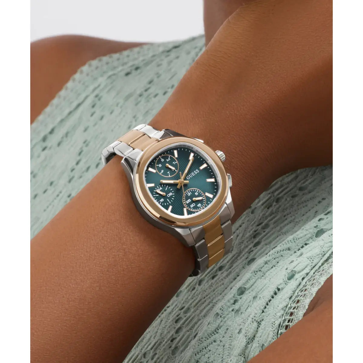 A two-tone stainless steel and gold-tone wristwatch with a dark green dial, worn on a person's wrist over a light green lace top.