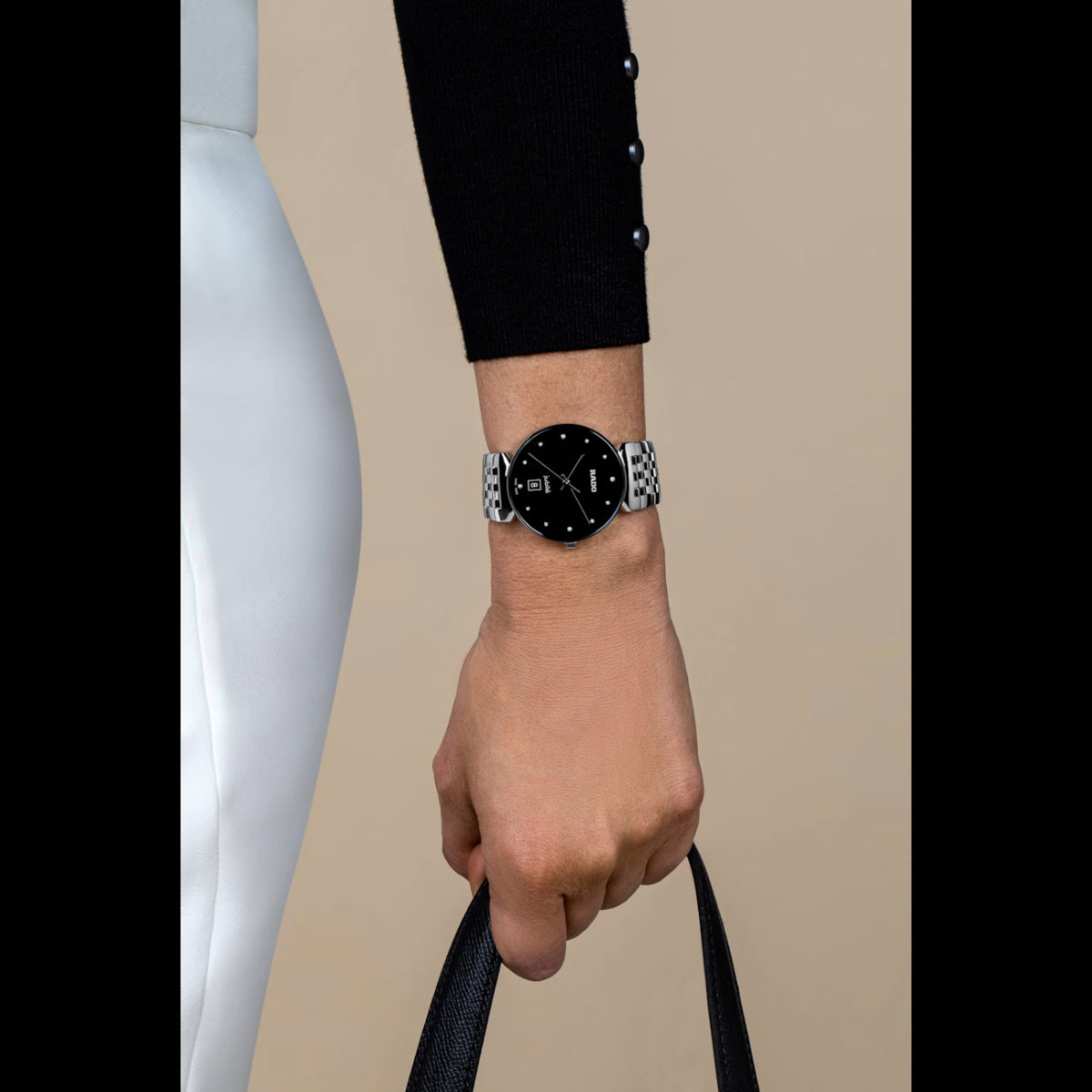 Hand wearing a watch with a black face and silver band, holding a black strap against a beige background.