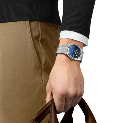 Hand wearing a silver watch with a blue face, holding a brown leather bag on a white background.