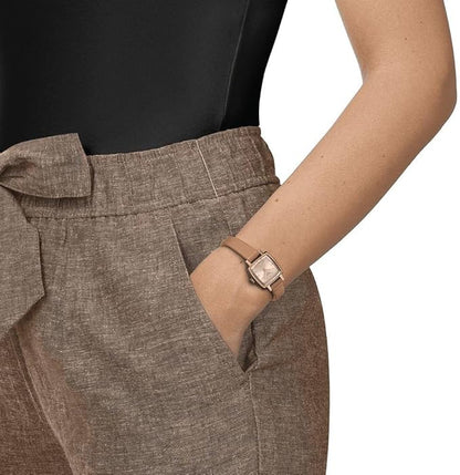 A woman's wrist wearing a silver Tissot Lovely Quartz Simple Analog Watch with a light brown leather strap, displayed against a backdrop of a black top and brown trousers.