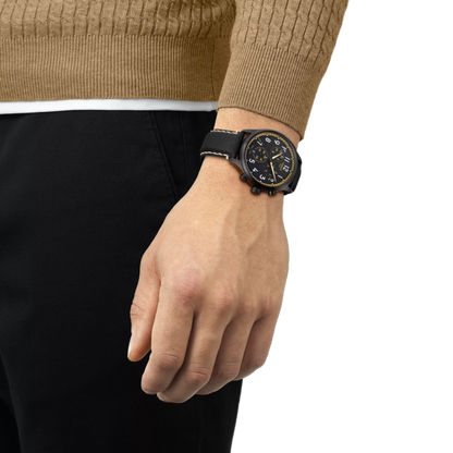 Hand wearing a black wristwatch with a detailed face, wearing a brown sweater and black pants on a white background.